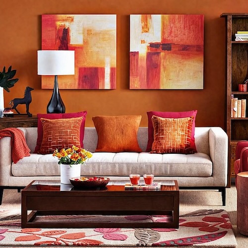 7 Cores Que Combinam Com Vermelho na Decoração Decoração de sala com parede laranja e sofá bege, almofadas coloridas em vermelho e laranja, quadros abstratos e mesa de centro de madeira